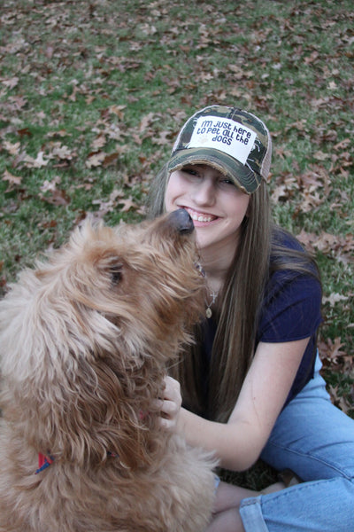 Woman wearing an I'm Just Here to Pet All the Dogs hat by BohoGroove with black I'm Just Here to Pet All the Dogs lettering and paw print graphic on a distressed off-white patch on a camo vintage distressed snapback while sitting outdoors with a dog.