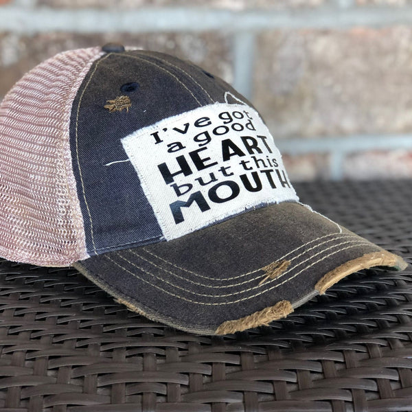 Left side view of I've Got a Good Heart, but This Mouth hat by BohoGroove with black lettering on a distressed off-white patch on a navy blue vintage distressed snapback trucker hat.