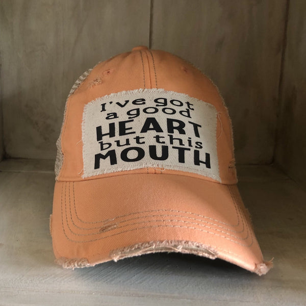 I've Got a Good Heart, but This Mouth hat by BohoGroove with black lettering on a distressed off-white patch on a sherbet vintage distressed snapback trucker hat.