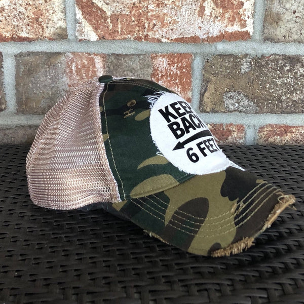 Left side view of the Keep Back 6 Feet hat by BohoGroove with black lettering and arrow graphic on a distressed off-white patch on a camo vintage distressed snapback trucker hat.