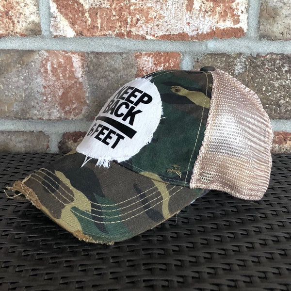 Right side view of the Keep Back 6 Feet hat by BohoGroove with black lettering and arrow graphic on a distressed off-white patch on a camo vintage distressed snapback trucker hat.