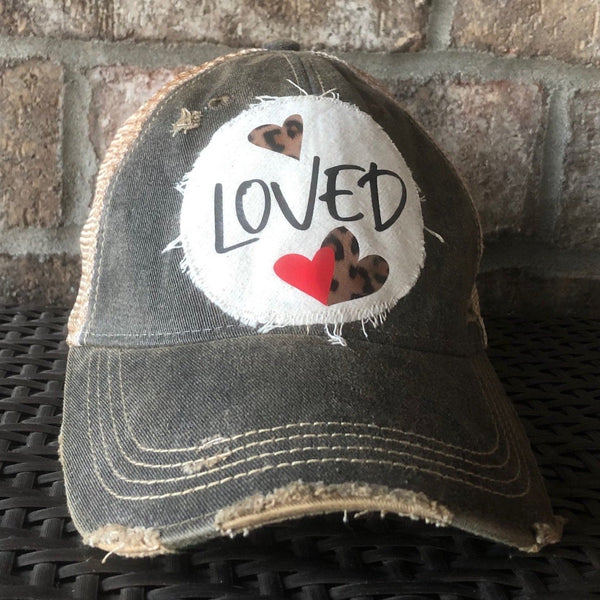 Loved hat by BohoGroove with black Loved lettering, red heart graphic, and leopard print heart graphics on a distressed off-white patch on a black vintage distressed snapback trucker hat.