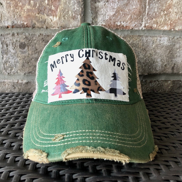 Merry Christmas Trees hat by BohoGroove with black Merry Christmas lettering and three Christmas tree graphics featuring red plaid, leopard print, and black buffalo plaid designs on a distressed off-white custom patch on a green vintage distressed snapback.