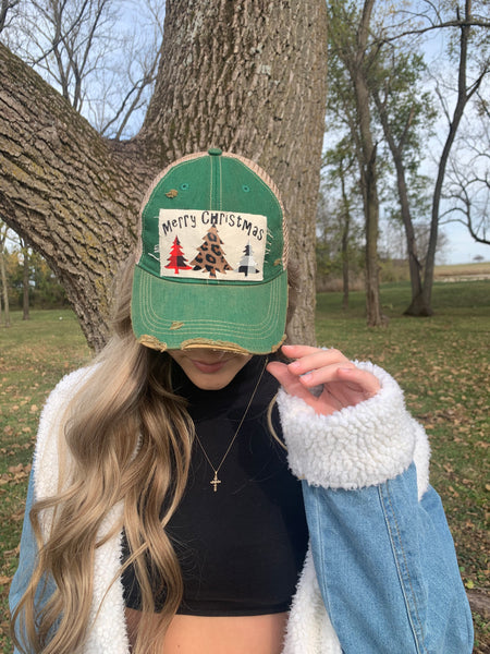 Merry Christmas Trees hat by BohoGroove with black Merry Christmas lettering and three Christmas tree graphics featuring pink plaid, leopard print, and black buffalo plaid designs on a distressed off-white custom patch on a green vintage distressed snapback worn by model.