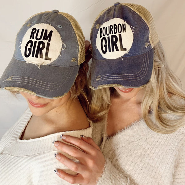 Two women wearing BohoGroove vintage distressed snapback trucker hats featuring distressed off-white patches with bold black Rum Girl and Bourbon Girl lettering.