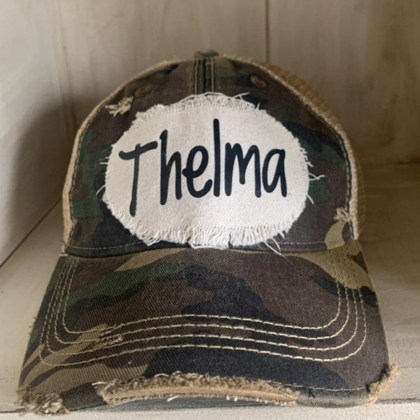 Thelma hat by BohoGroove with black Thelma lettering on a distressed off-white patch on a camo vintage distressed snapback trucker hat.